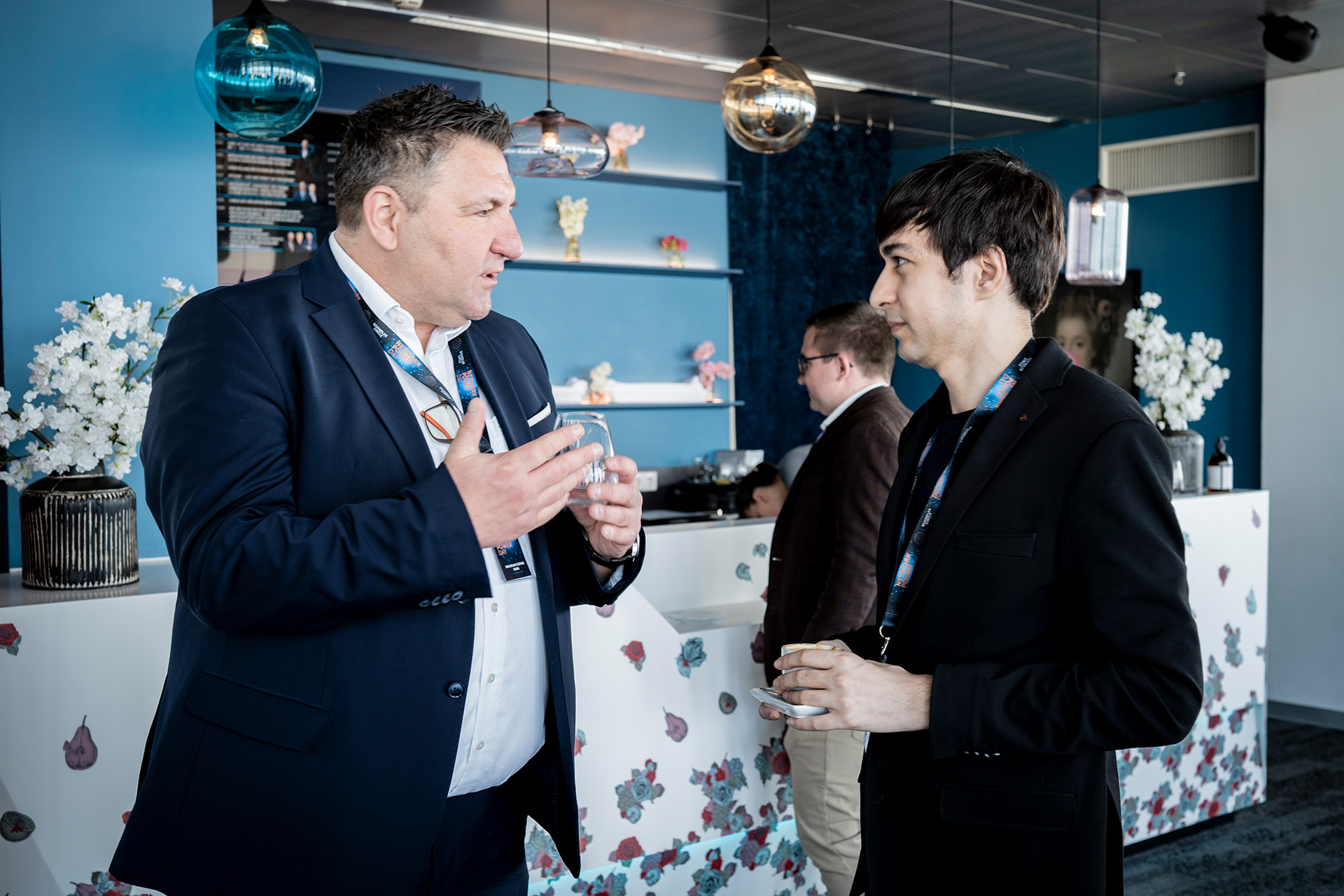 Two men in suits engaged in conversation at a conference, surrounded by attendees and presentation materials.