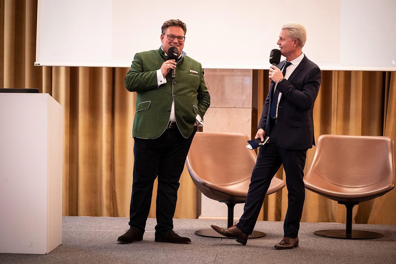 Three men seated in chairs, engaged in discussion at a conference setting.