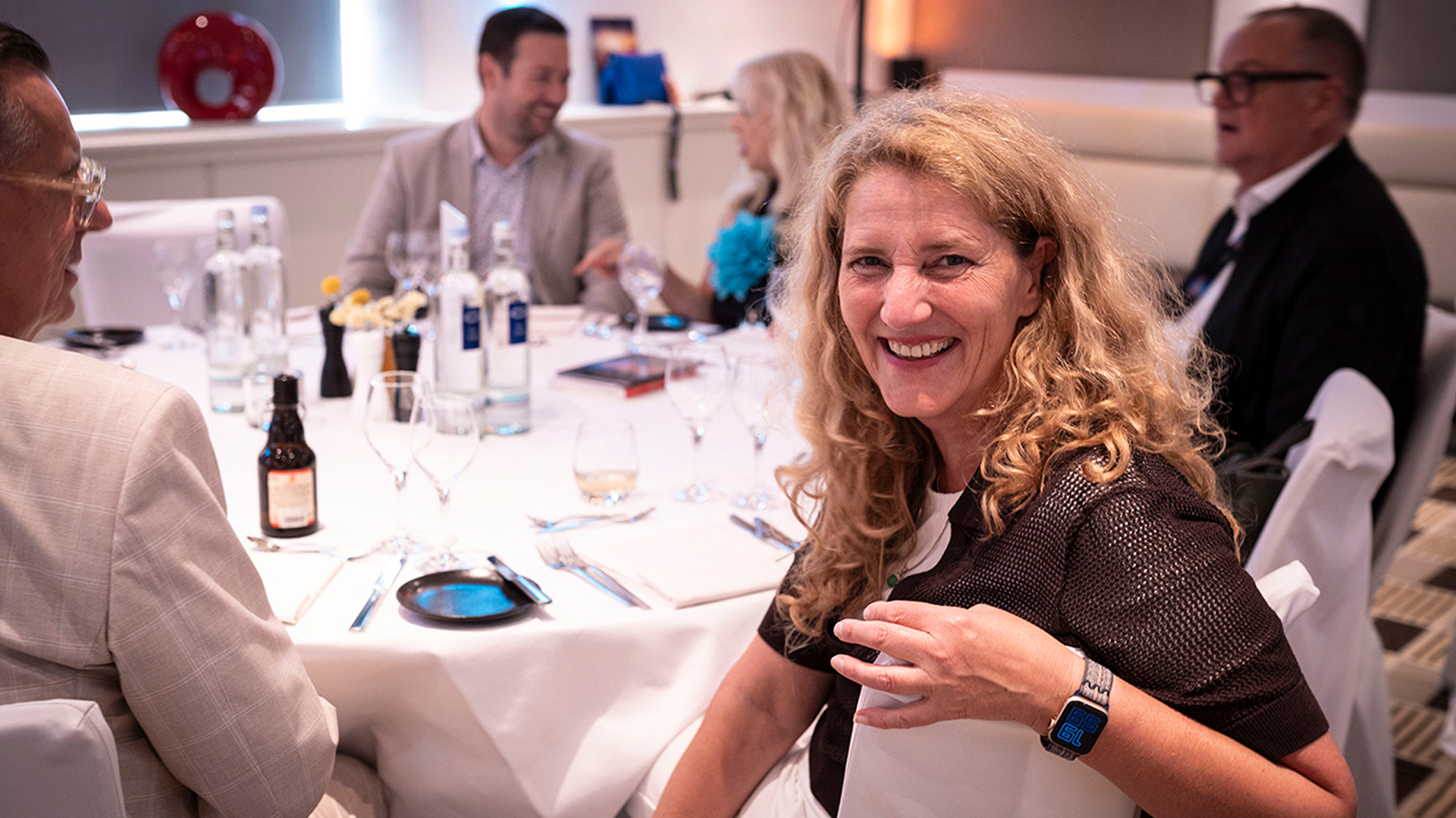 Smiling woman with curly hair at a formal dinner table, wearing a smartwatch. Background shows people in conversation, wine glasses, and bottled water.