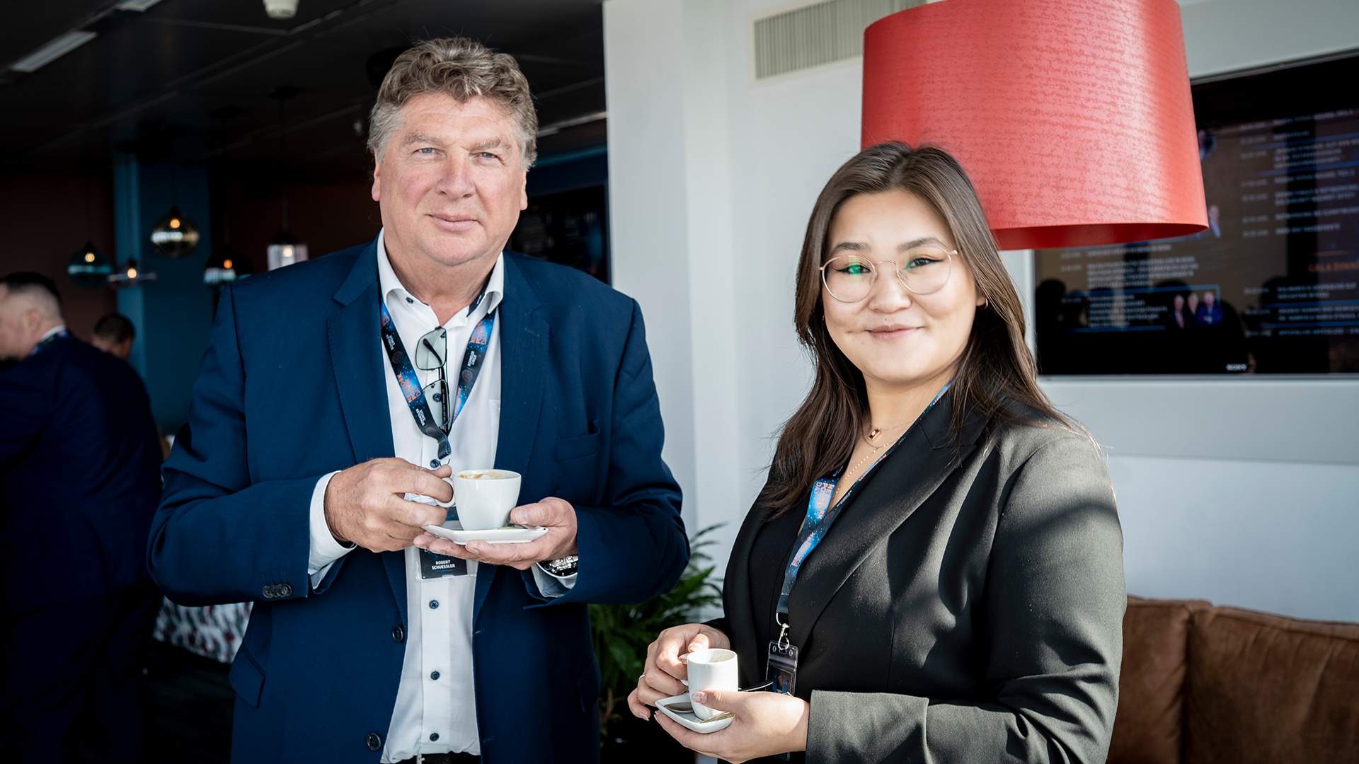 Two people in business attire are smiling while holding espresso cups in a modern lounge. A red lamp and additional people in the background.