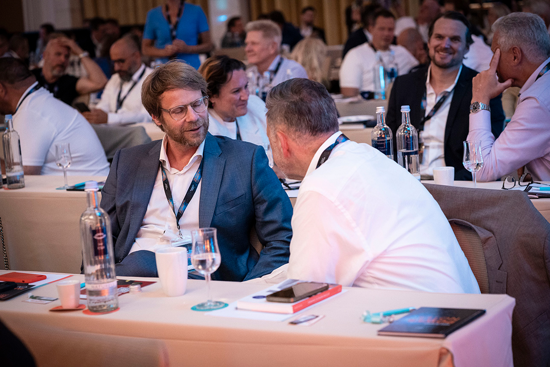 A conference setting with attendees seated at tables. Two men in the foreground engage in conversation, surrounded by water bottles, notebooks, and lanyards. The mood is professional and collaborative.
