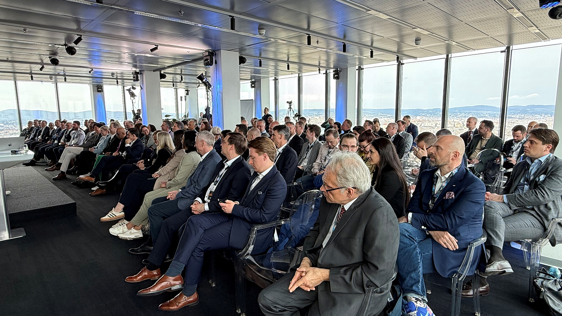 Audience in formal attire attentively seated in a modern, glass-walled conference room with city views, conveying a professional and engaged atmosphere.
